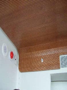 Surfacing Solution | Curved tambour wood ceiling by Surfacing Solution with vent, red sensor, and two white fixtures above a white wall in modern interior