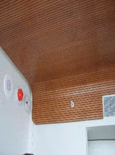 Surfacing Solution | Curved tambour wood ceiling by Surfacing Solution with vent, red sensor, and two white fixtures above a white wall in modern interior