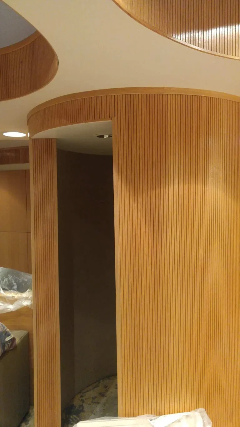 Surfacing Solution | Curved tambour wood panels with vertical grooves create a modern room divider, featuring a dark interior and ceiling lights behind